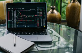 Laptop displaying cryptocurrency stocks and graphs on a glass table with a notepad.